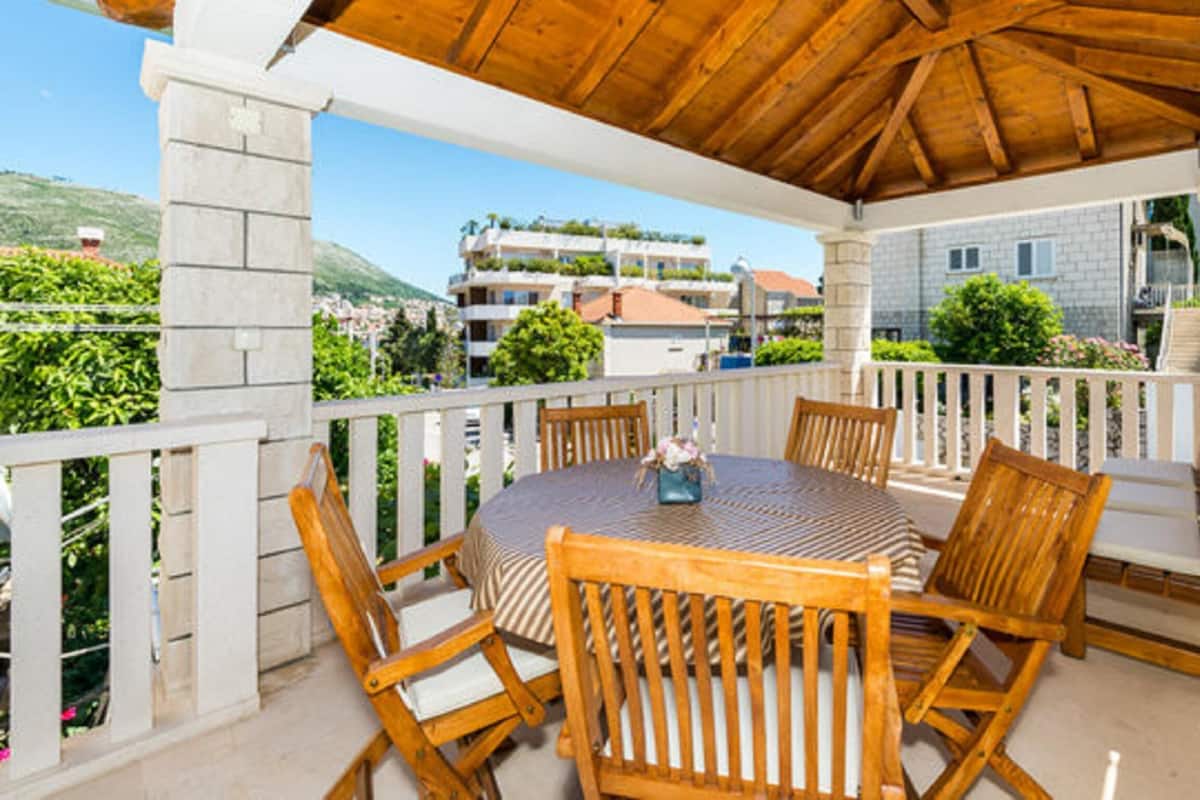 A covered outdoor seating area with a round table and wooden chairs, overlooking a residential area with houses and greenery.