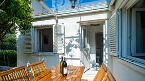 A wooden table with four chairs and a bottle of wine, set on a patio with a white wall and shutters.
