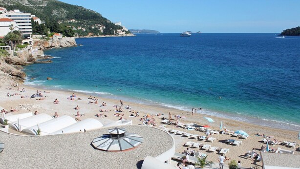 A beach with sun loungers and umbrellas, a clear blue sea, and buildings in the background.