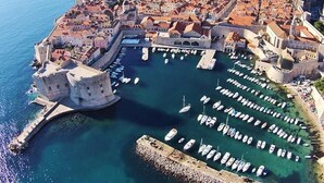 An aerial view of a coastal city with historic architecture, a harbor with numerous boats, and a fortified castle-like structure.