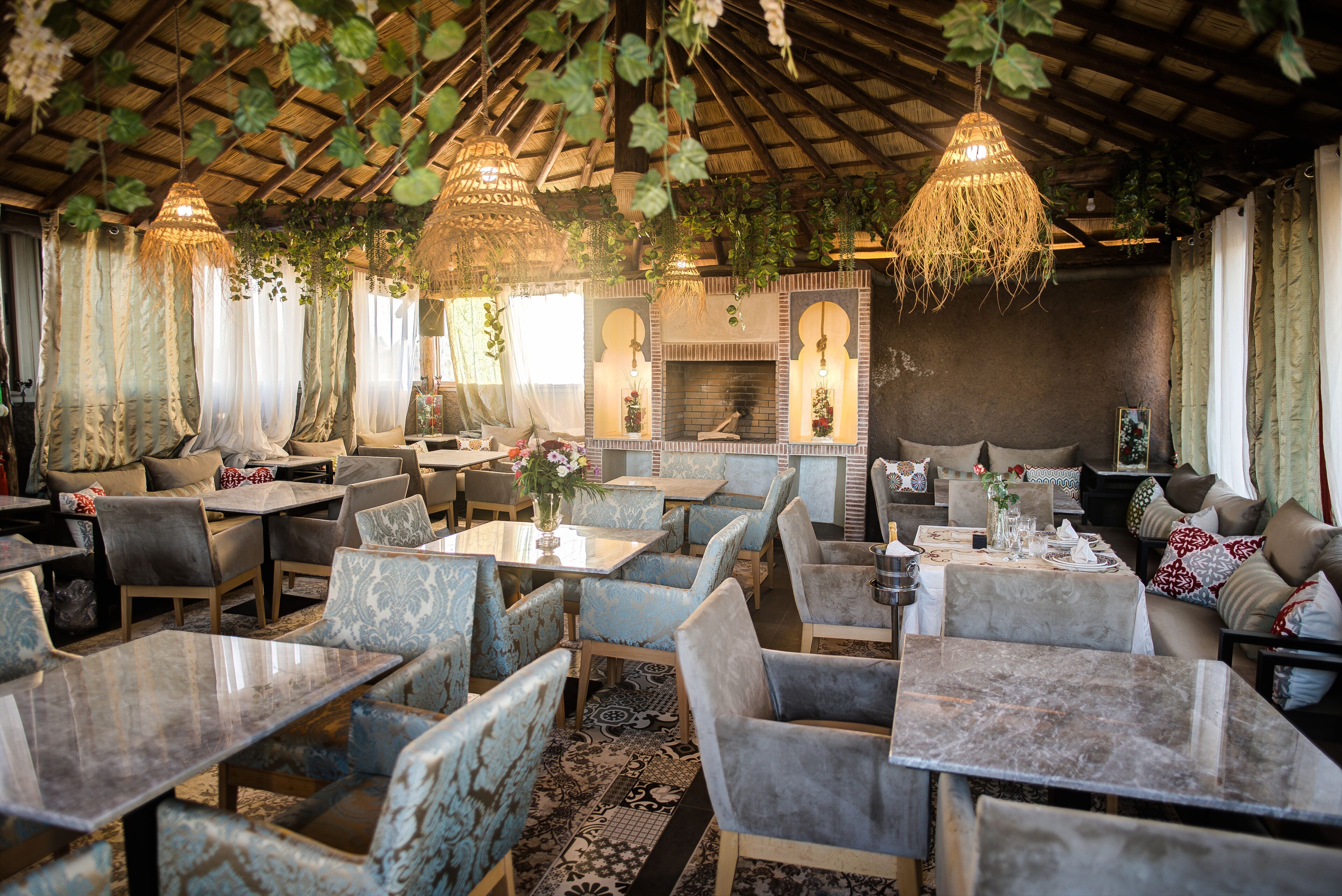 A restaurant with a rustic interior, featuring hanging plants, a brick fireplace, and a variety of seating arrangements including tables and chairs.