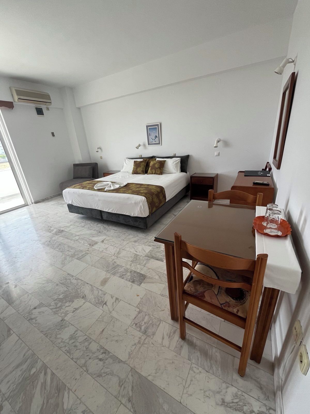 Une chambre d’hôtel avec un lit, un bureau et une chaise. Il y a un tableau au mur et un appareil de climatisation au plafond.