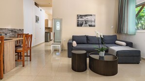 A modern living room with a sofa, two round side tables, a dining area with a wooden table and chairs, and a kitchen in the background. - Vlycha Beach Studios & Apartments (Rhodes)
