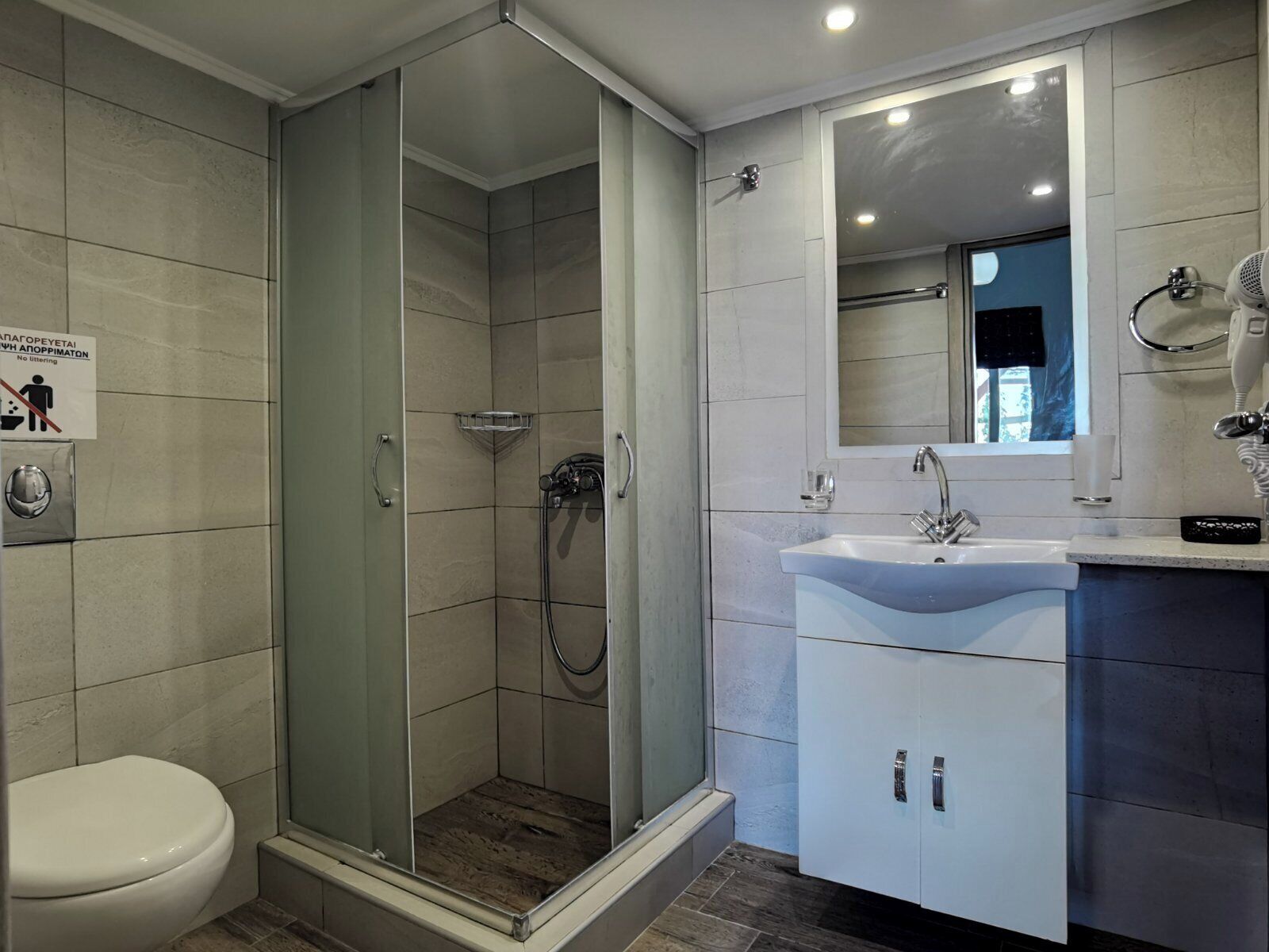Egy modern fürőszoba üvegfalas zuhanyzóval, fehér mosdókagylóval és WC-vel.