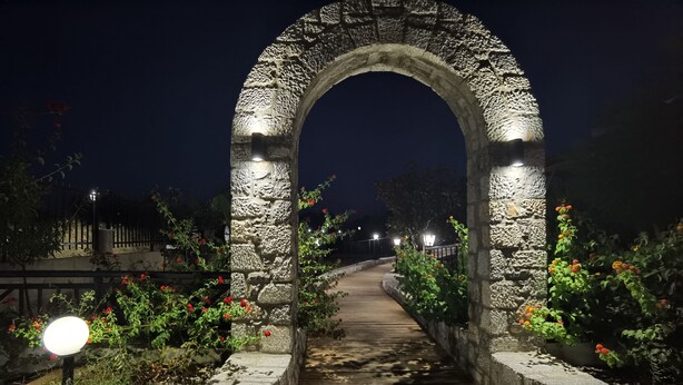 A stone archway with a wooden pathway leading through it, surrounded by flowering plants and illuminated by a single light source. - Vlycha Beach Studios & Apartments (Rhodes)