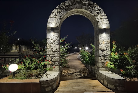 A stone archway with a wooden pathway leading through it, surrounded by flowering plants and illuminated by a single light source.