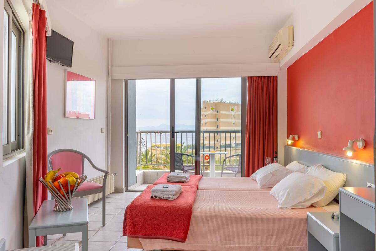 Ein Hotelzimmer mit zwei Betten, einem Sessel, einem kleinen Tisch mit Obstschale und einem Balkon mit Blick auf Gebäude.