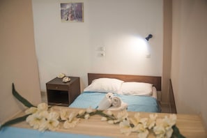 Ein ordentlich bezogenes Bett mit weißen Kissen und einer blauen Tagesdecke, ein kleiner Nachttisch mit Blumen in einer Vase und eine wandmontierte Leuchte.