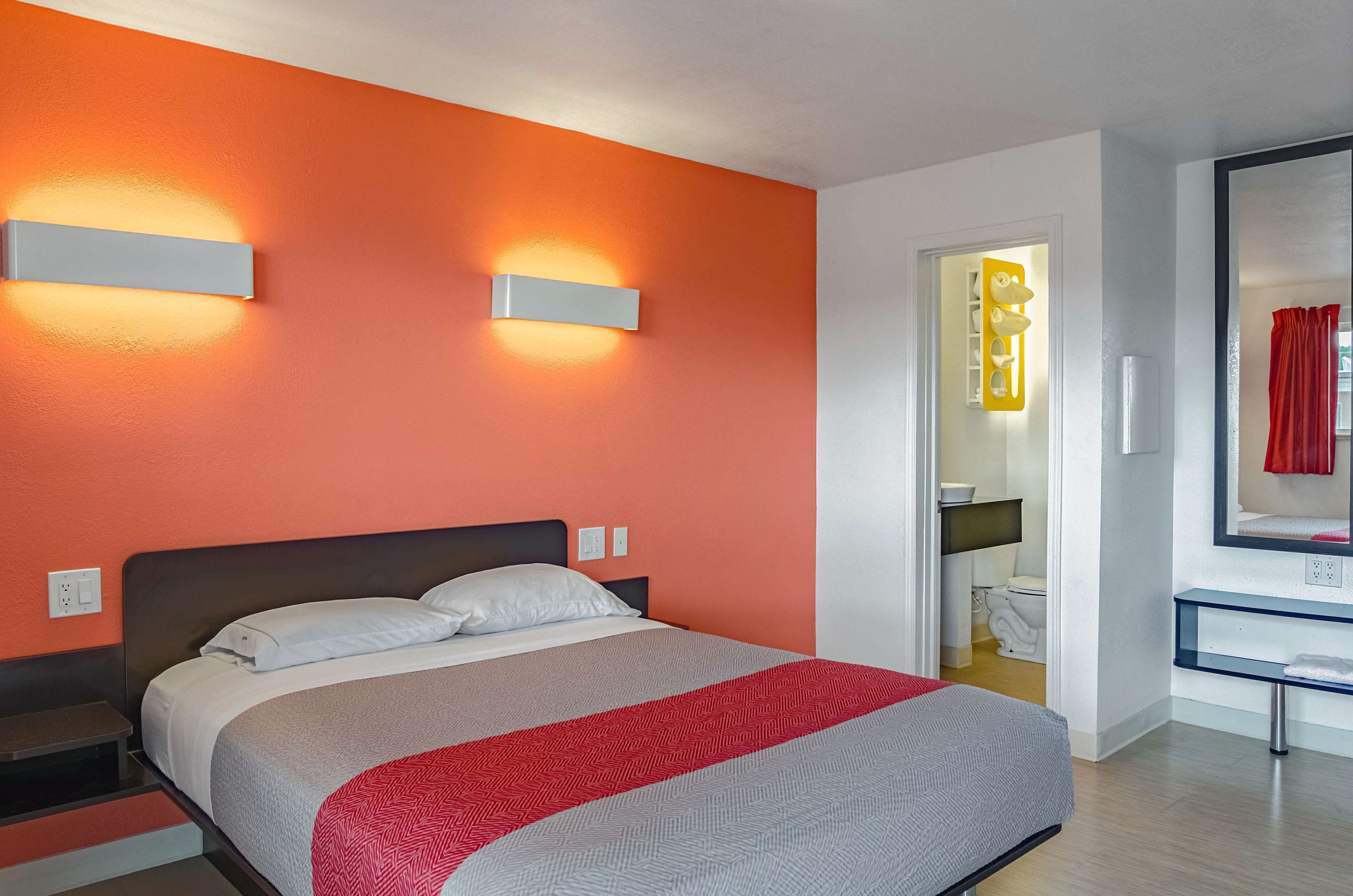 Habitación de hotel con cama, mesita de noche, espejo y baño visible a través de una puerta abierta.