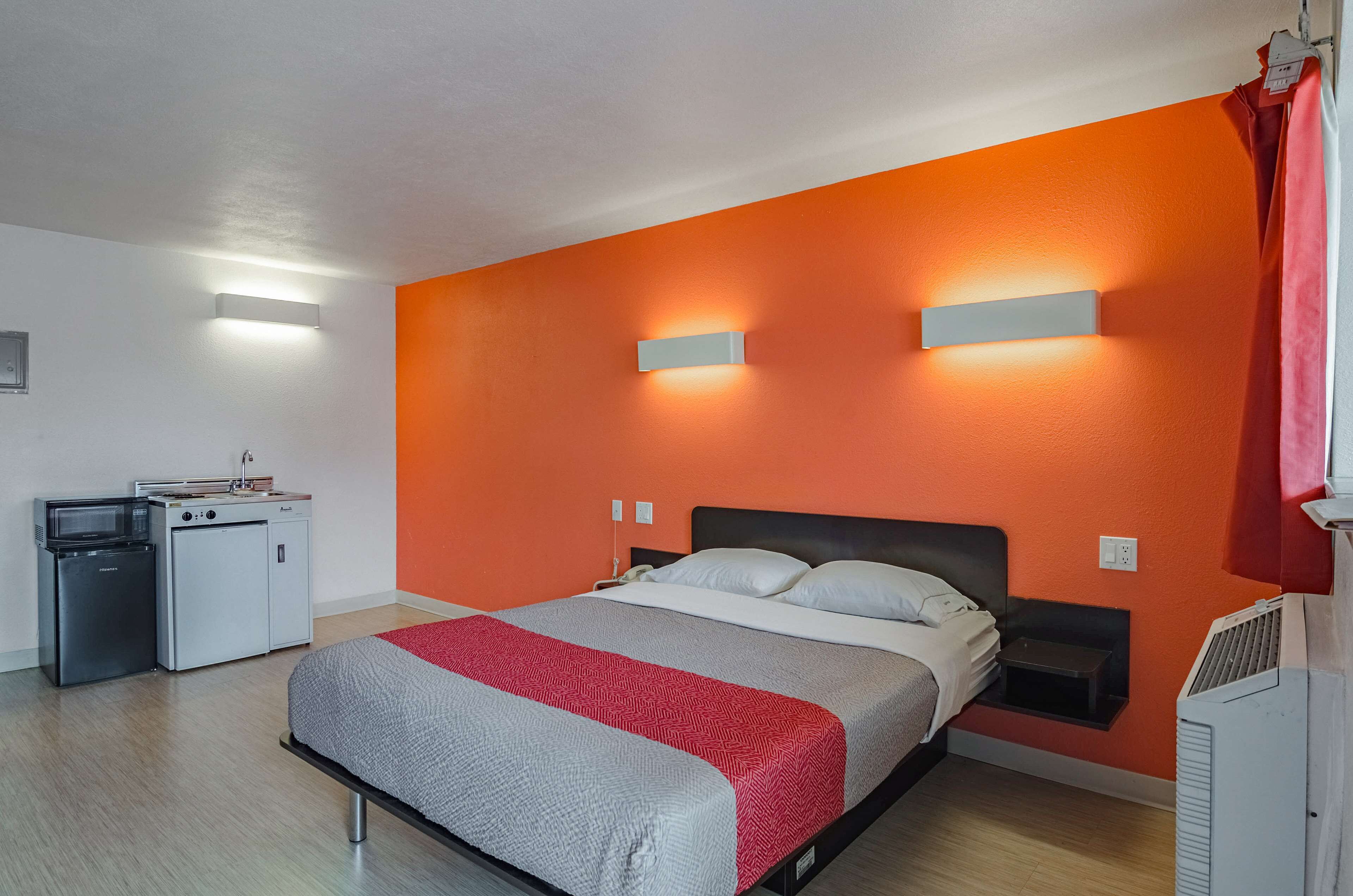A room with a bed, a small kitchenette, and a bright orange wall.