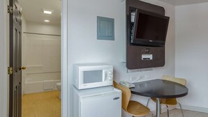 Ein kompaktes Zimmer mit einer weißen Mikrowelle auf einem kleinen, weißen Kühlschrank, einem flachbildfernseher an der Wand, einem Holzstuhl und einem kleinen, runden Tisch.