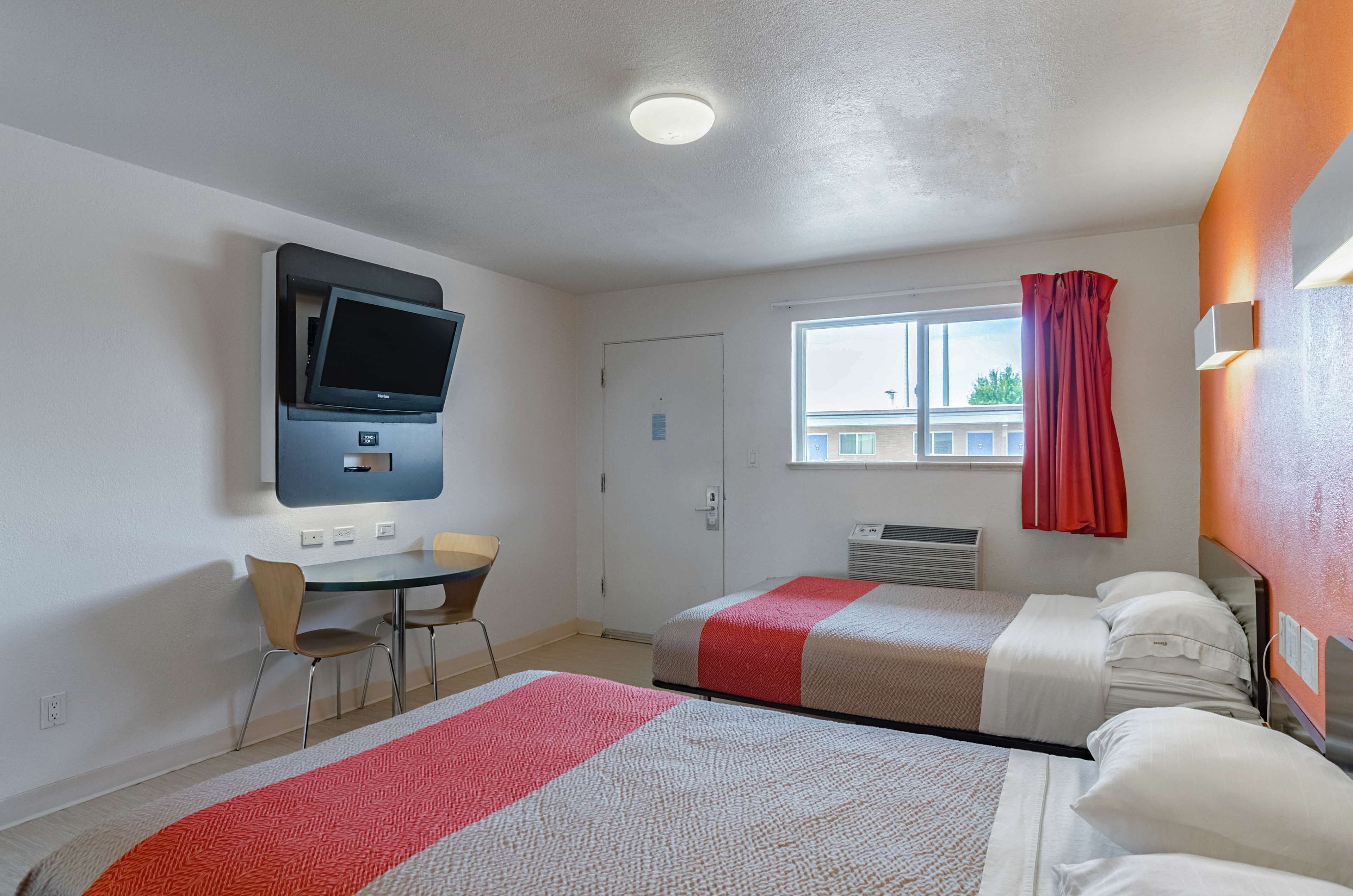 Ein Hotelzimmer mit zwei Betten, einem Schreibtisch mit Stuhl, einem an der Wand montierten Fernseher und einem Fenster mit Vorhang.