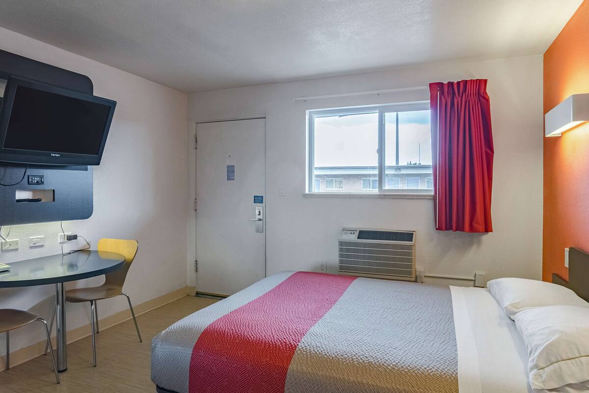 Ein Hotelzimmer mit Bett, Schreibtisch mit Stuhl, Fernseher, Fenster mit Vorhängen und einer Klimaanlage.