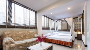 A hotel room with a bed, a sofa, a small table with a vase, and large windows with blinds.