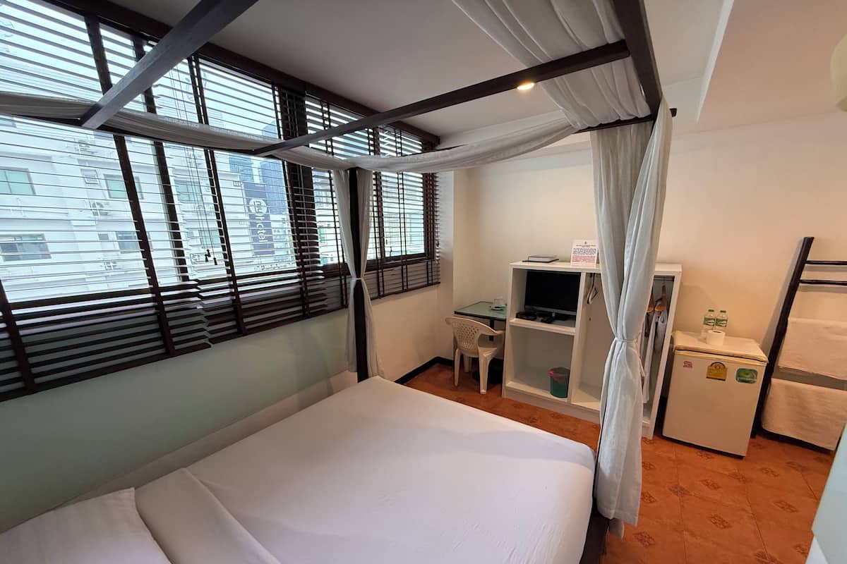 A hotel room with a bed, a desk, a chair, a small refrigerator, and a window with blinds.