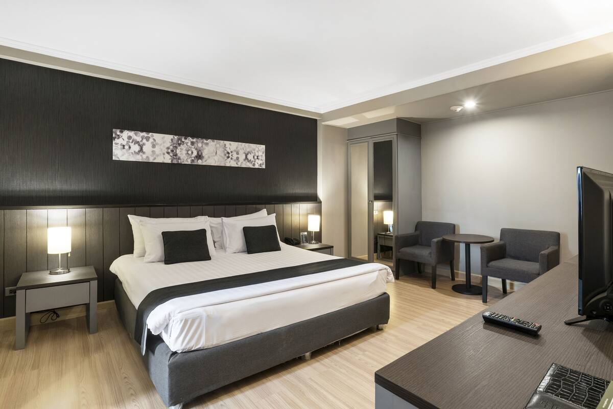 A modern hotel room with a large bed, bedside tables with lamps, a desk with a chair, a television, and a mirror.