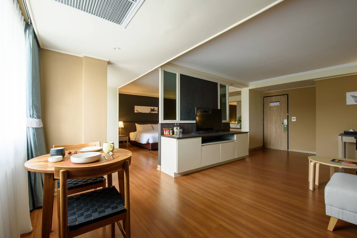 A modern hotel room with a wooden dining table, a sofa, a TV, and a kitchenette.