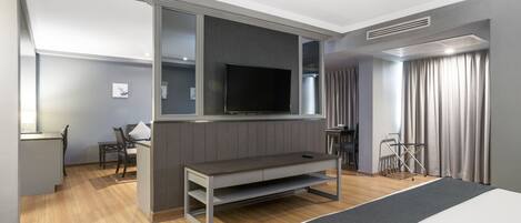 Habitació d'hotel moderna amb un llit gran, un televisió de pantalla plana penjada a la paret, una zona de seients petita amb taula i cadires, i una finestra amb cortines.