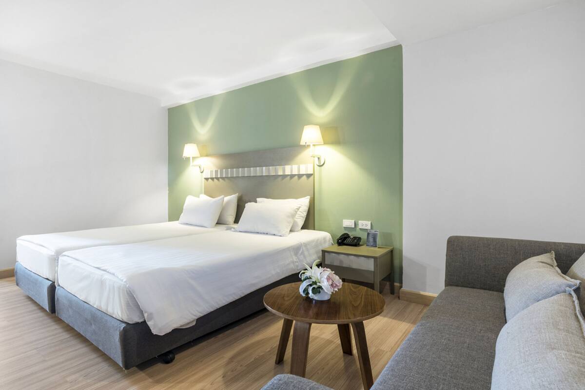 A hotel room with a large bed, a bedside table with a lamp, a small round table with a vase, and a sofa.