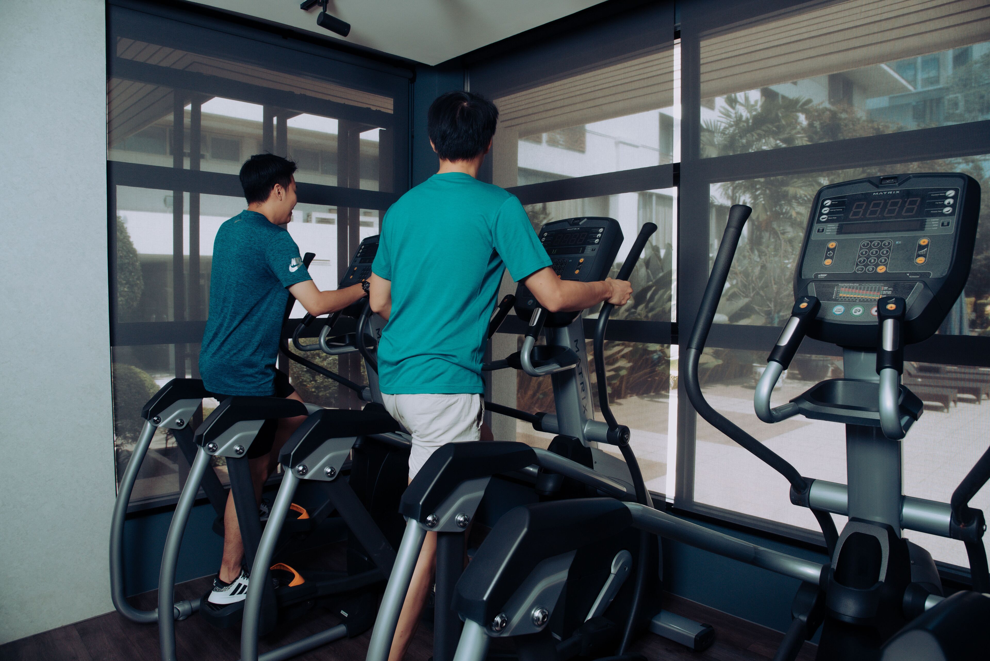 Deux personnes font de l'exercice sur des tapis de course dans une salle de sport dotée de grandes fenêtres.
