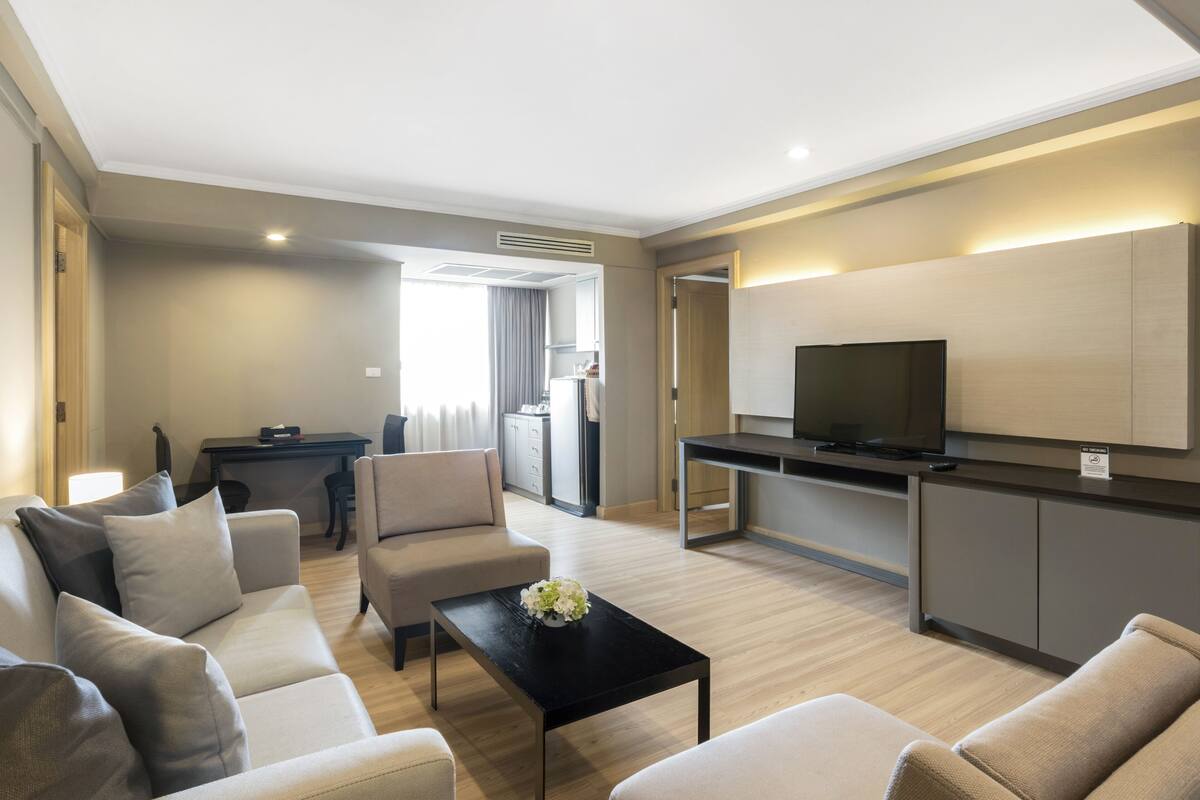 A modern hotel room with a sofa, armchairs, a coffee table, a TV, and a kitchenette.