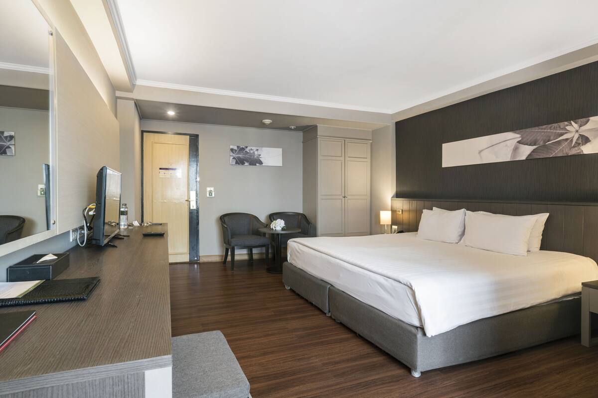 A modern hotel room with a large bed, a desk with a computer, a chair, and a small table with a vase.