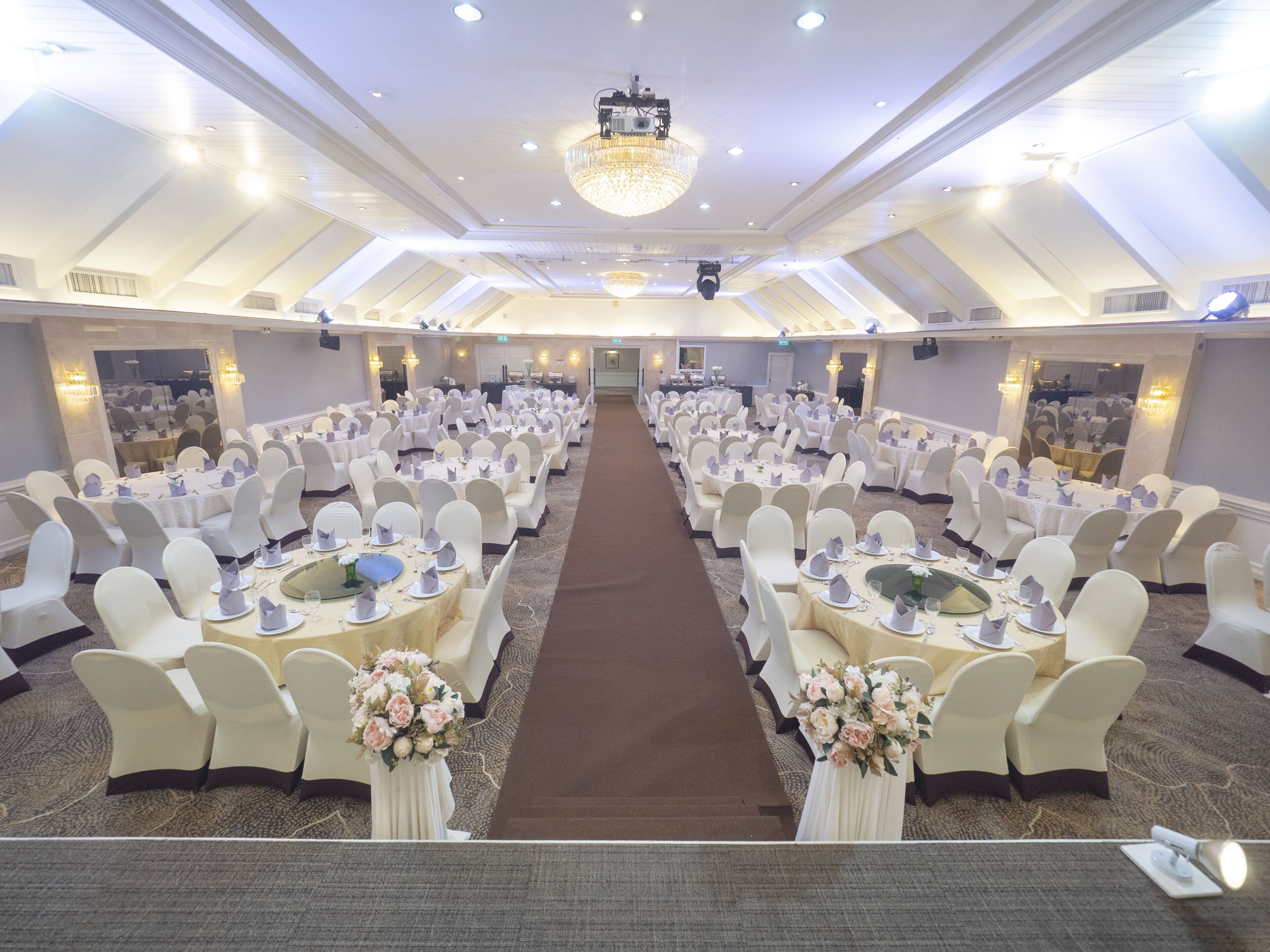 A large banquet hall with round tables and white chairs, a central aisle, and a chandelier.