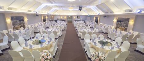 A large banquet hall with round tables and white chairs, a central aisle, and a chandelier.