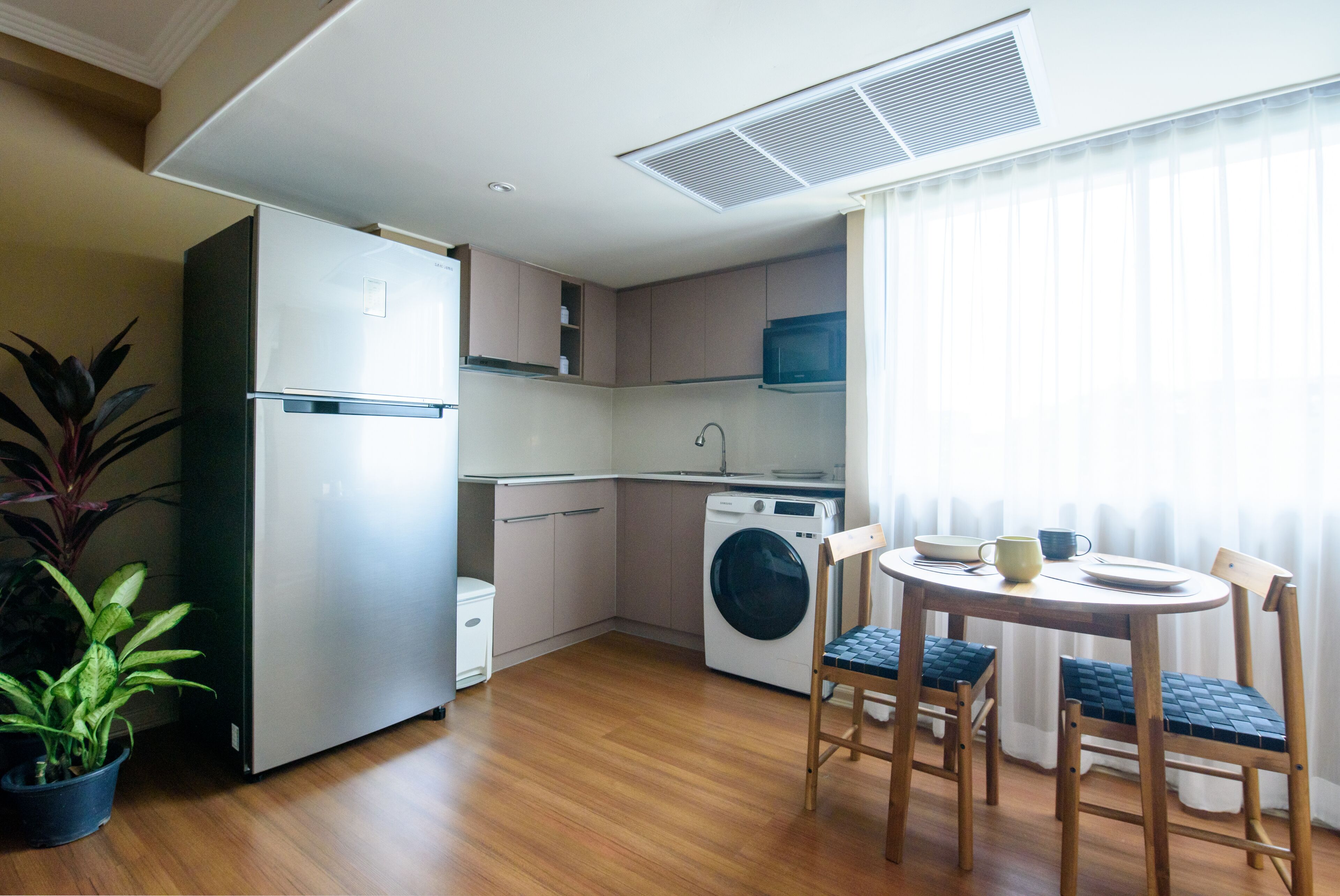 A compact kitchen with a refrigerator, washing machine, and microwave. There is a dining table with chairs and a potted plant.