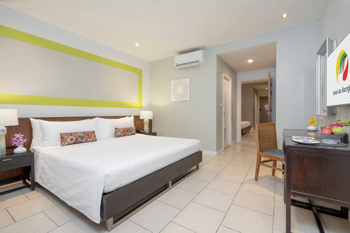 Une chambre d’hôtel avec un grand lit, des tables de chevet, un bureau avec des fruits et une vue sur le couloir.