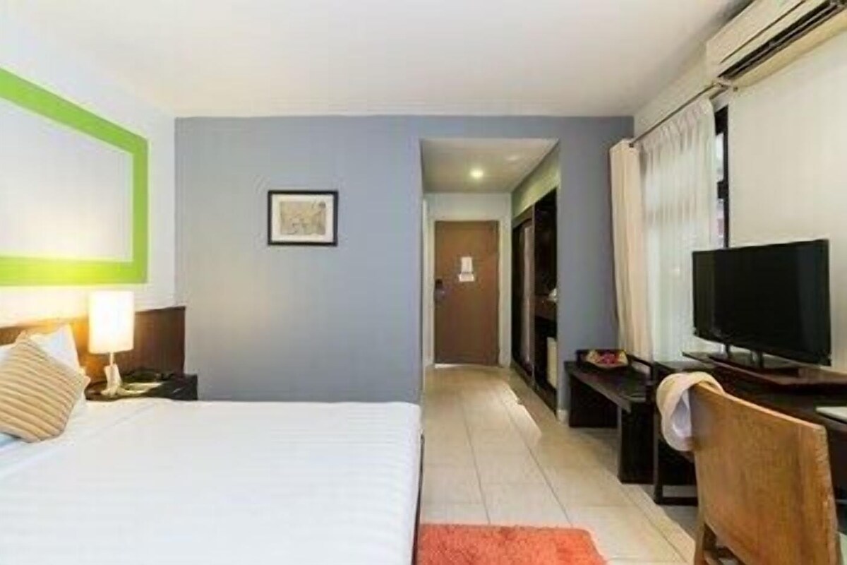 Ein Hotelzimmer mit einem Bett, einem Schreibtisch mit Computer, einem Fernseher, einem gerahmten Bild an der Wand und einer Tür zu einem anderen Raum.
