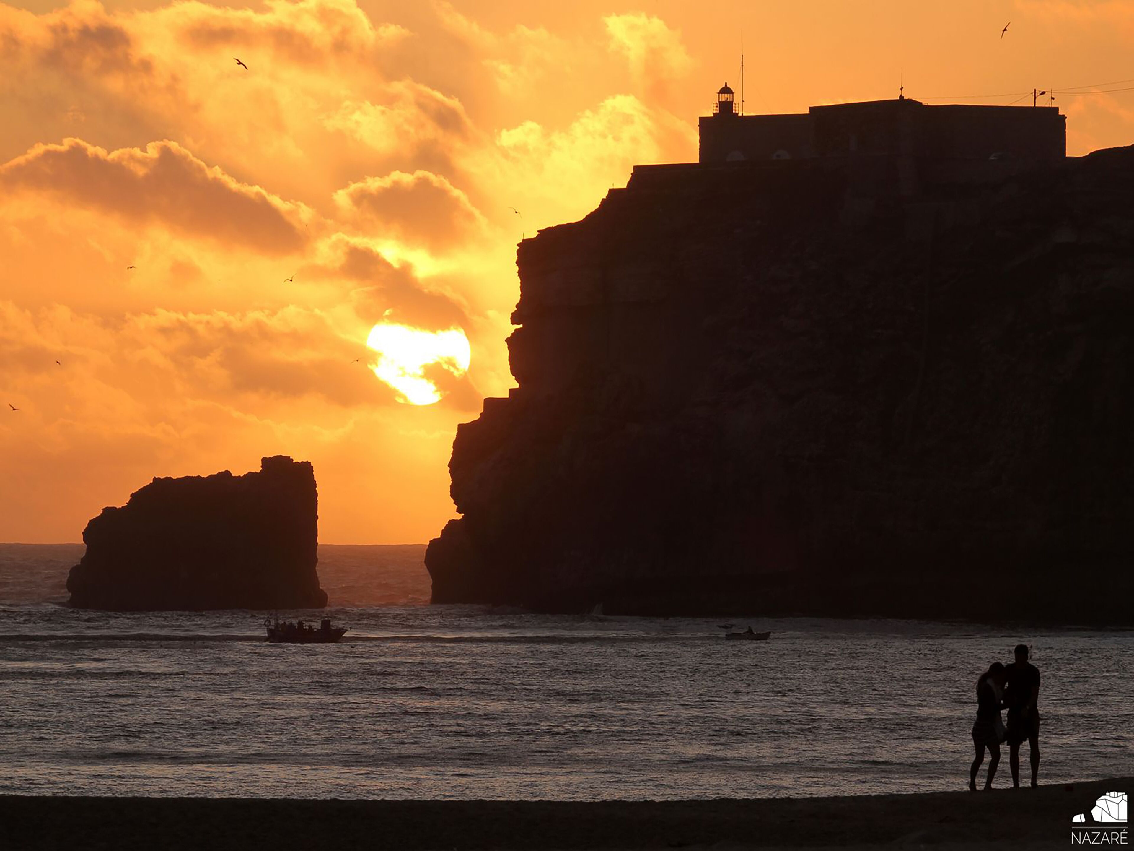 A coastal sunset with cliffs, a lighthouse, and a couple by the water.