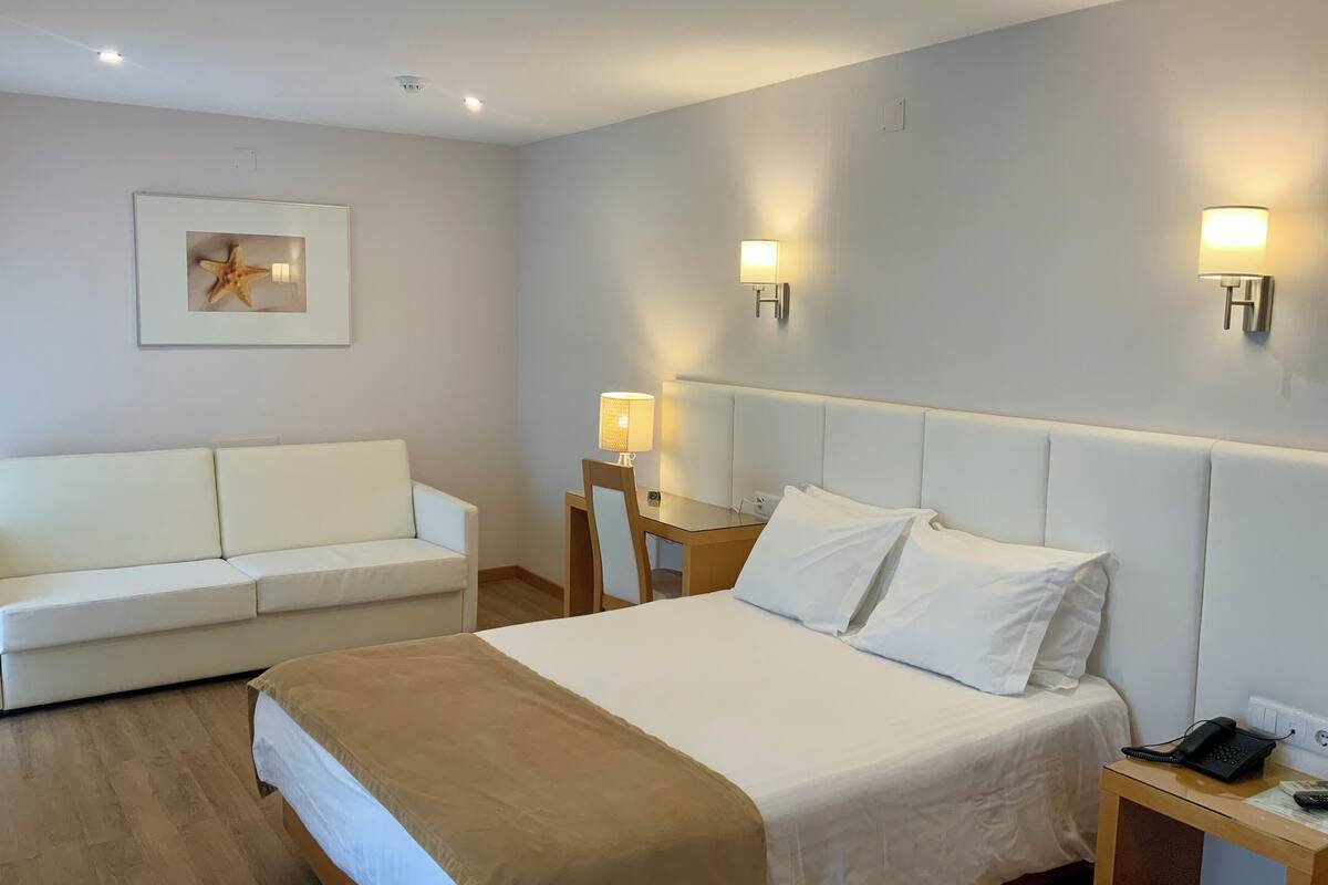 Habitación de hotel con cama, sofá blanco, mesita de noche con lámpara y aplique de pared.
