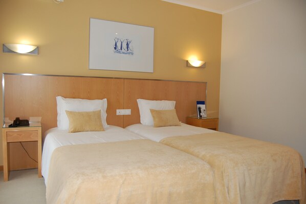 Quarto de hotel com duas camas, cabeceira de madeira, luminárias laterais, mesa de cabeceira e uma obra de arte emoldurada na parede.