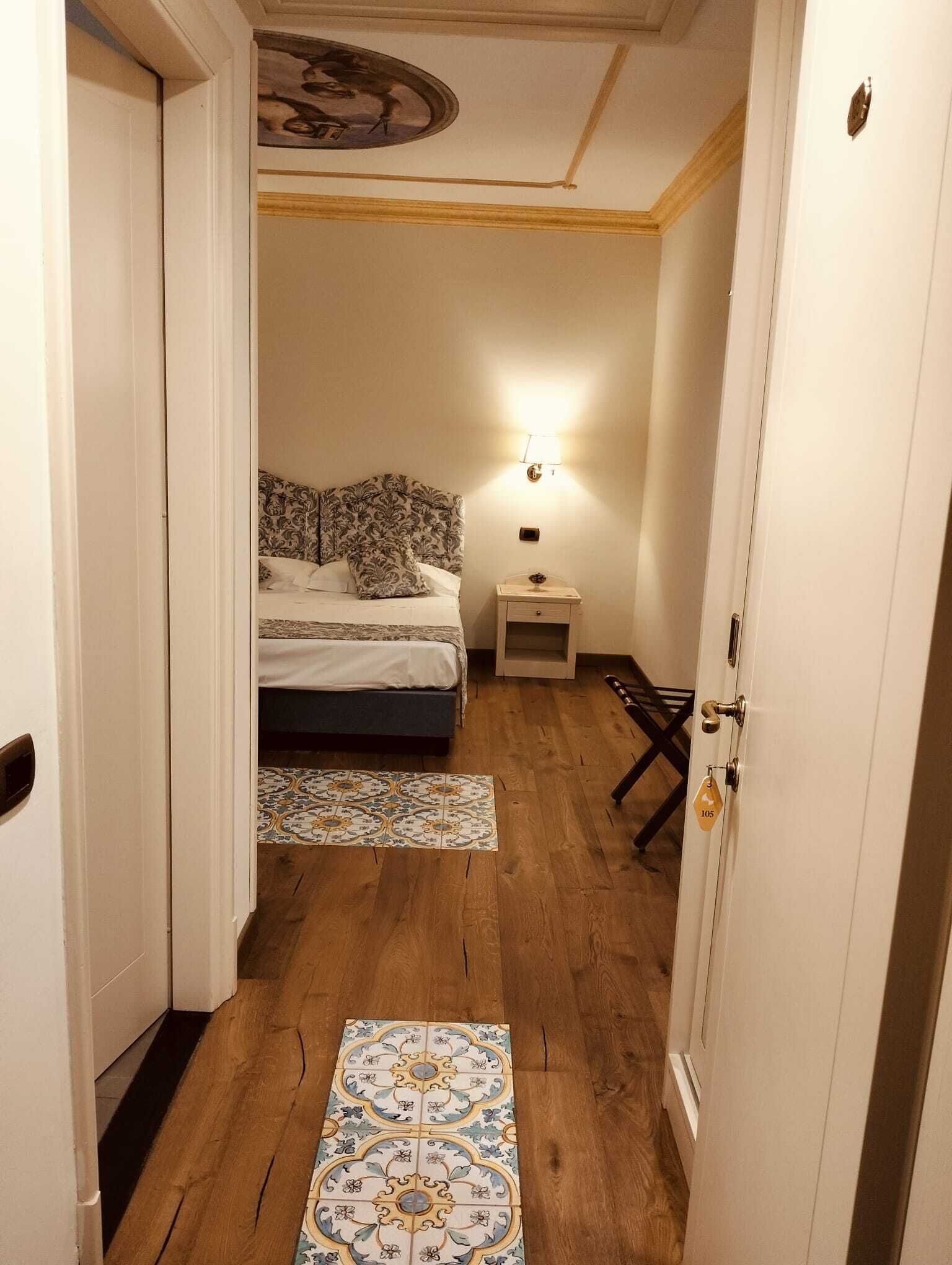 A narrow hallway leading to a bedroom with a bed, nightstand, and a wall-mounted lamp.