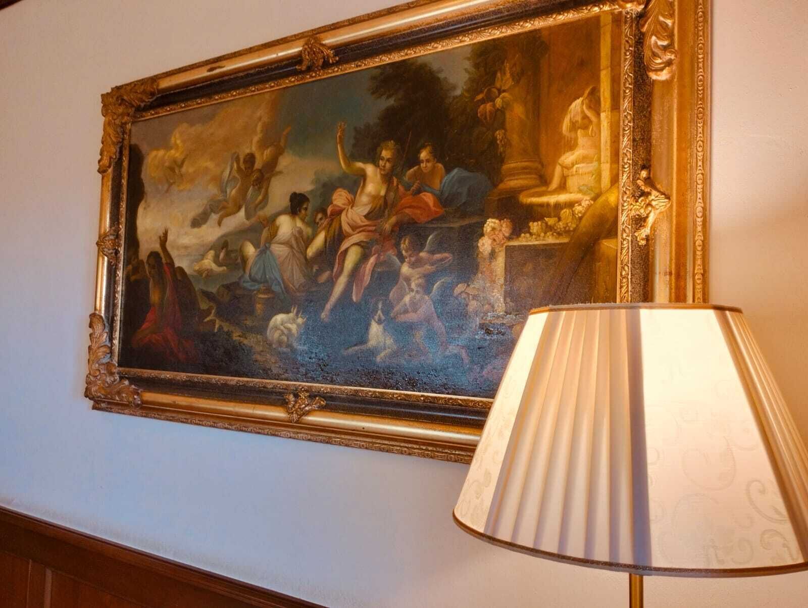En klassisk målning i en guldfärgad ram hänger på en vägg, bredvid en vit lampa med en veckad lampskärm.