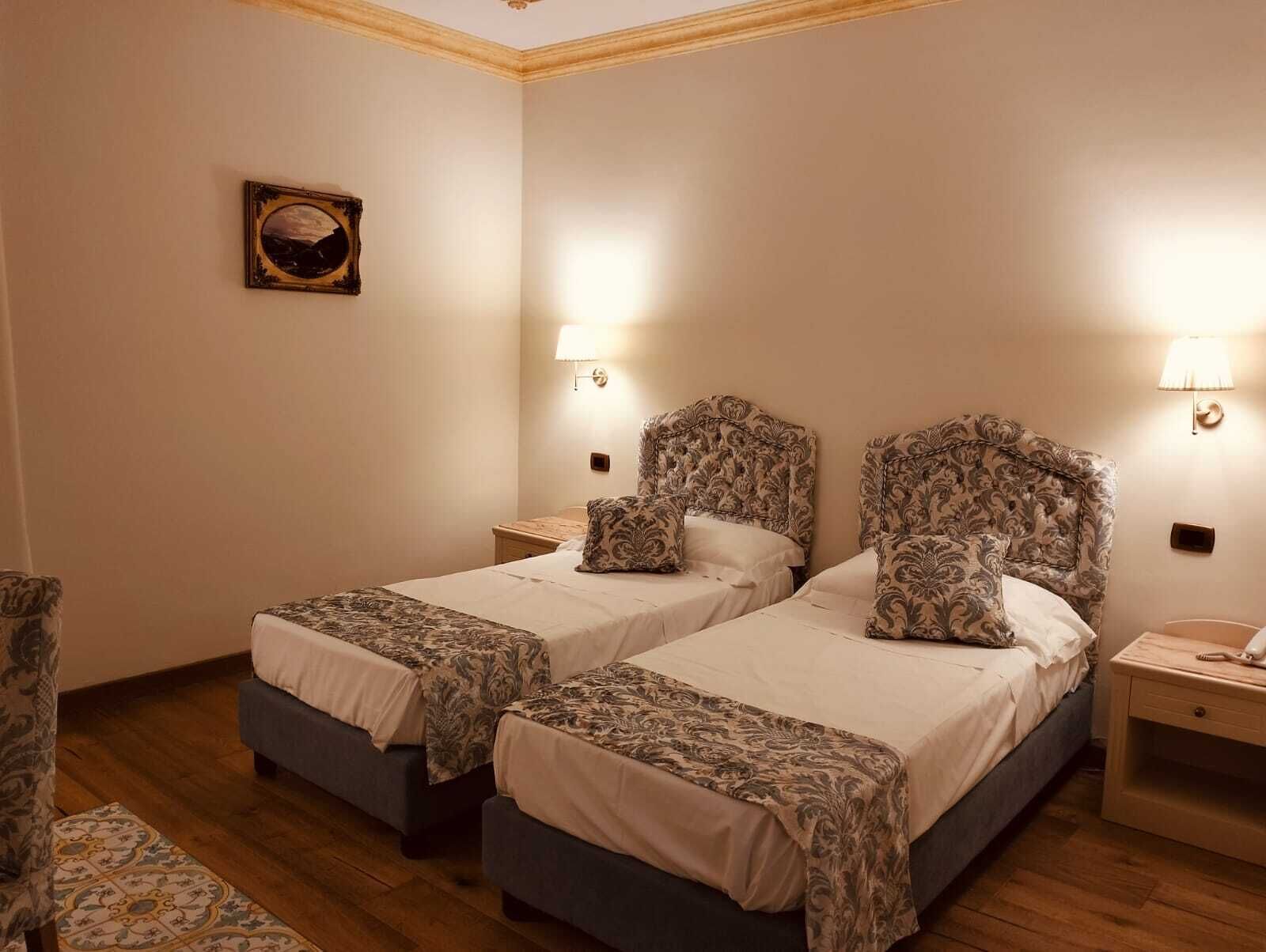 Habitación de hotel con dos camas, cada una con un cabecero estampado y almohadas a juego, una mesita de noche pequeña con lámpara, y un cuadro enmarcado en la pared.