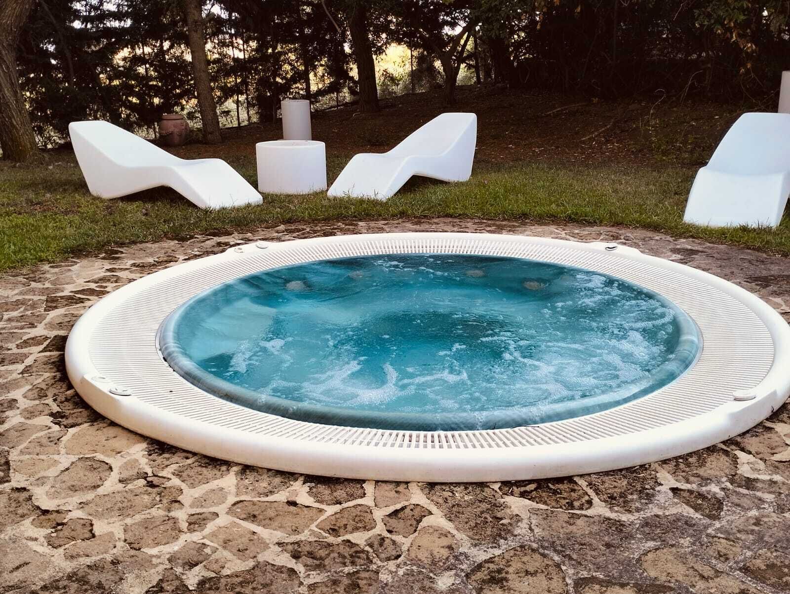 Un jacuzzi rodeado de baldosas de piedra, con sillones blancos para relajarse y un área verde de fondo.
