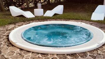 Un jacuzzi rodeado de baldosas de piedra, con sillones blancos para relajarse y un área verde de fondo.