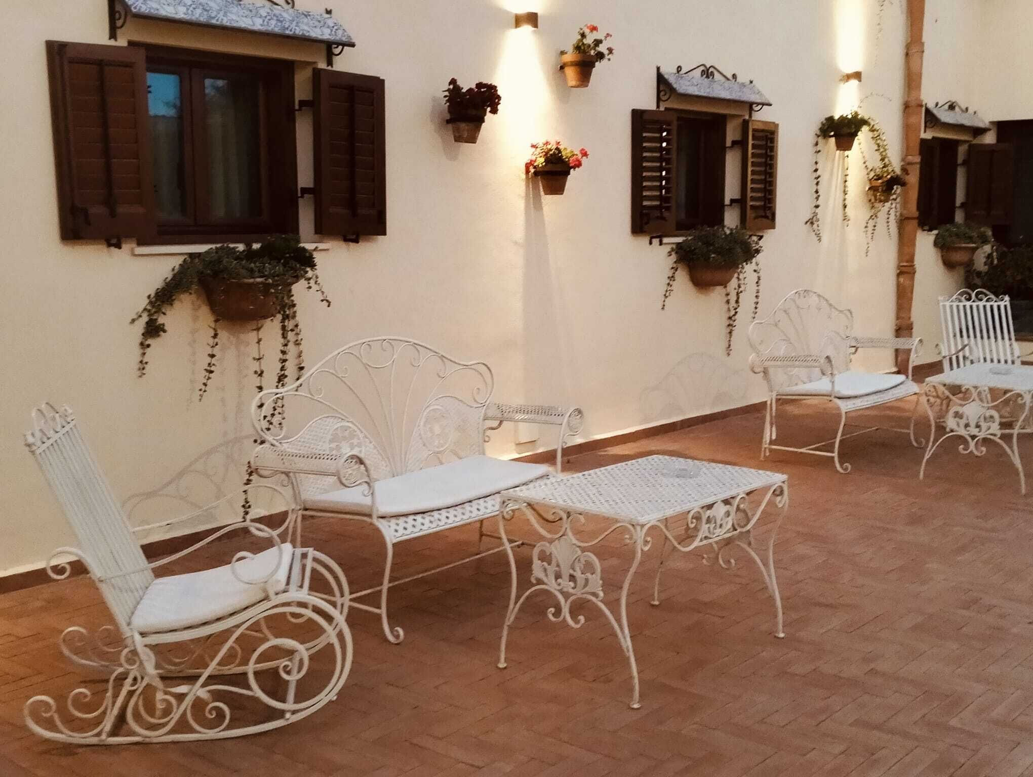 A corridor with white metal chairs and tables, wooden shutters on windows, and hanging planters with greenery.