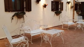 A corridor with white metal chairs and tables, wooden shutters on windows, and hanging planters with greenery.