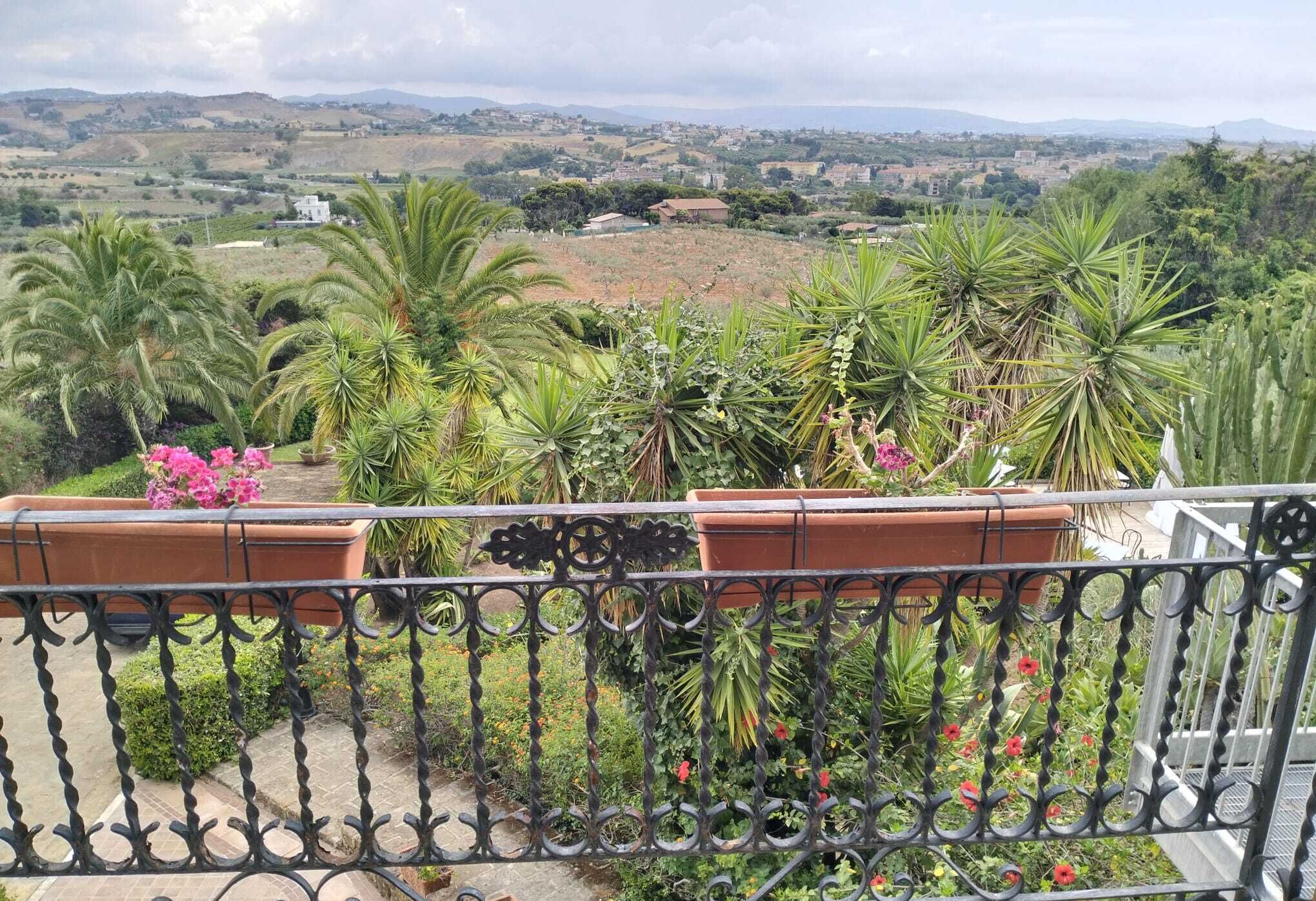 Un balcón con barandilla de hierro forjado que da a un jardín ajardinado con diversas plantas y flores.