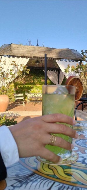 A person holding a glass with a lime slice, sitting at an outdoor table with a patterned tablecloth, under a thatched gazebo. - Baglio Della Luna (Agrigento)
