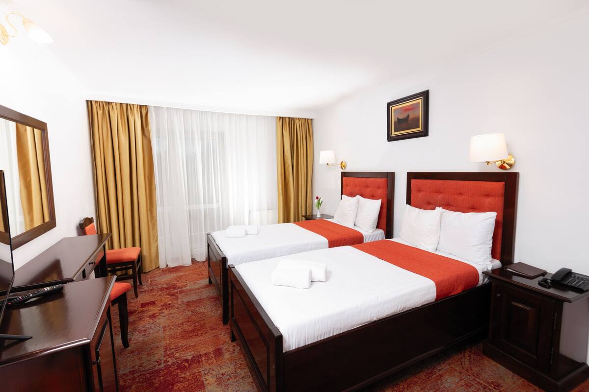 Une chambre d’hôtel avec deux lits, un bureau, une chaise, une lampe de bureau, une table de chevet et un tableau au mur.