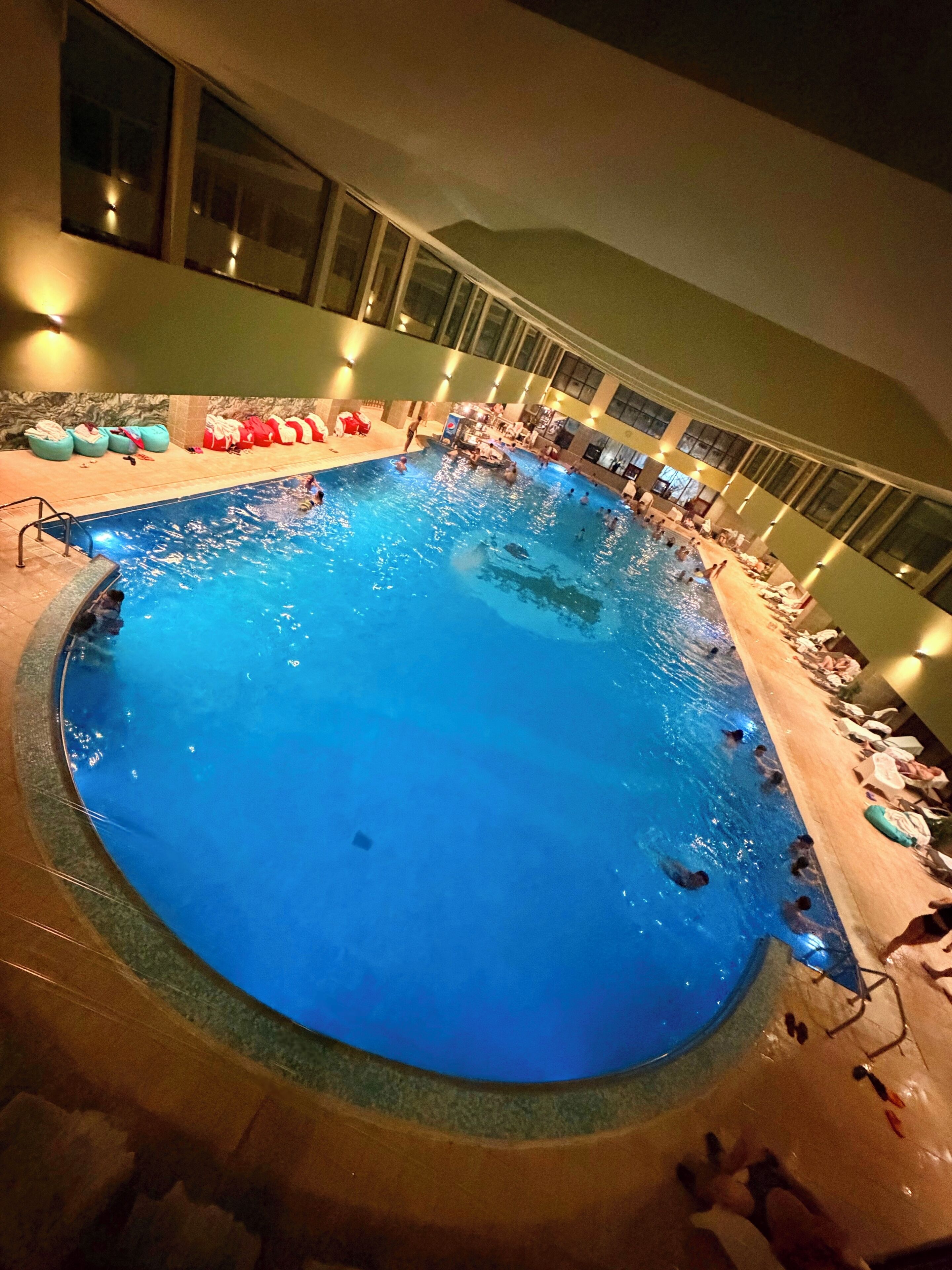 An indoor swimming pool with a tiled edge, a lounge area with chairs and towels, and a high ceiling with windows.