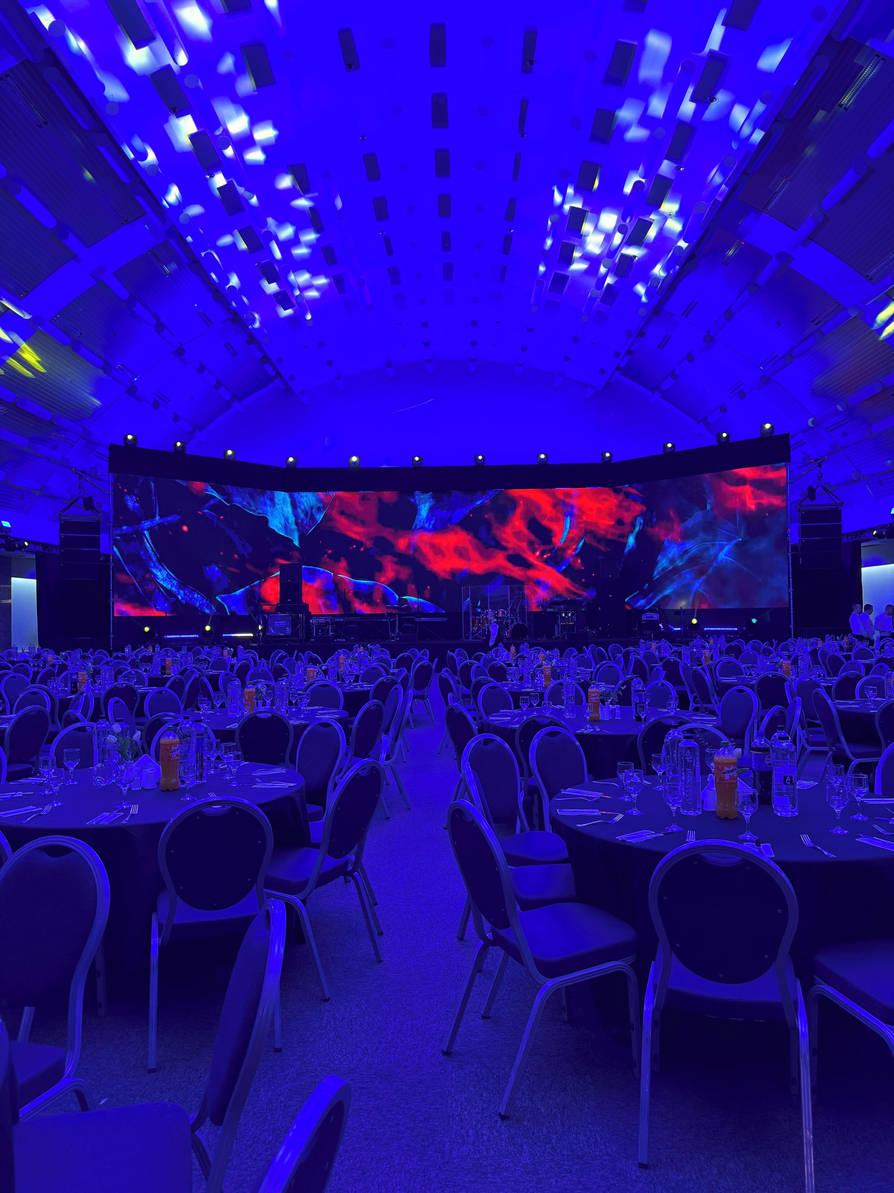 A large conference room with rows of tables and chairs, a large screen at the front, and a stage with lighting.