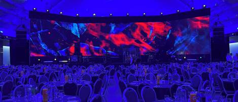 A large conference room with rows of tables and chairs, a large screen at the front, and a stage with lighting.