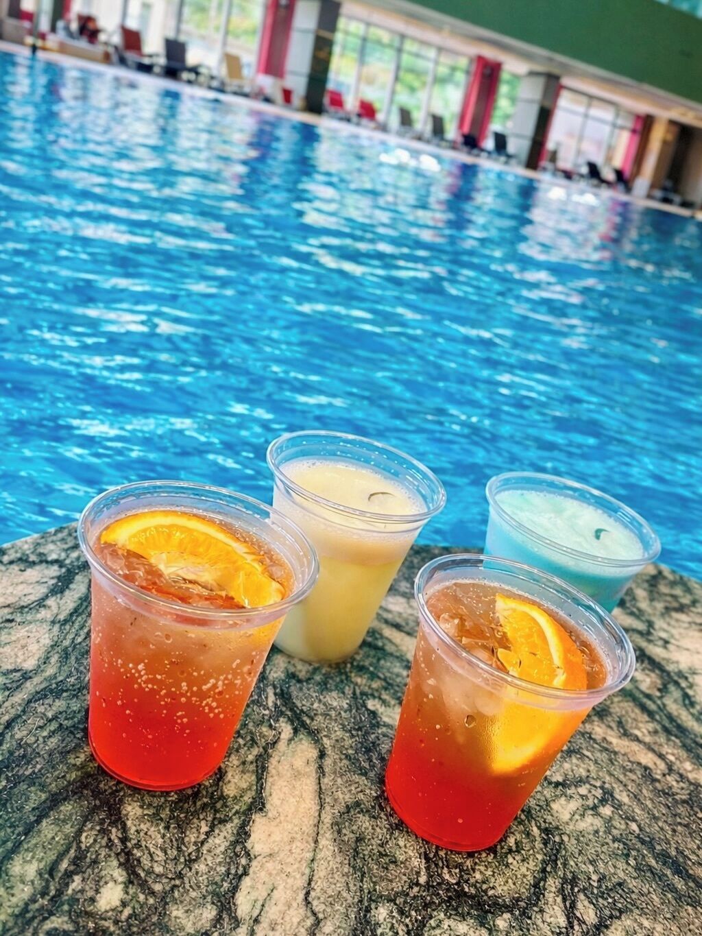 Escena junto a la piscina con tres bebidas en vasos transparentes, uno con una rodaja de naranja, y una piscina al fondo.