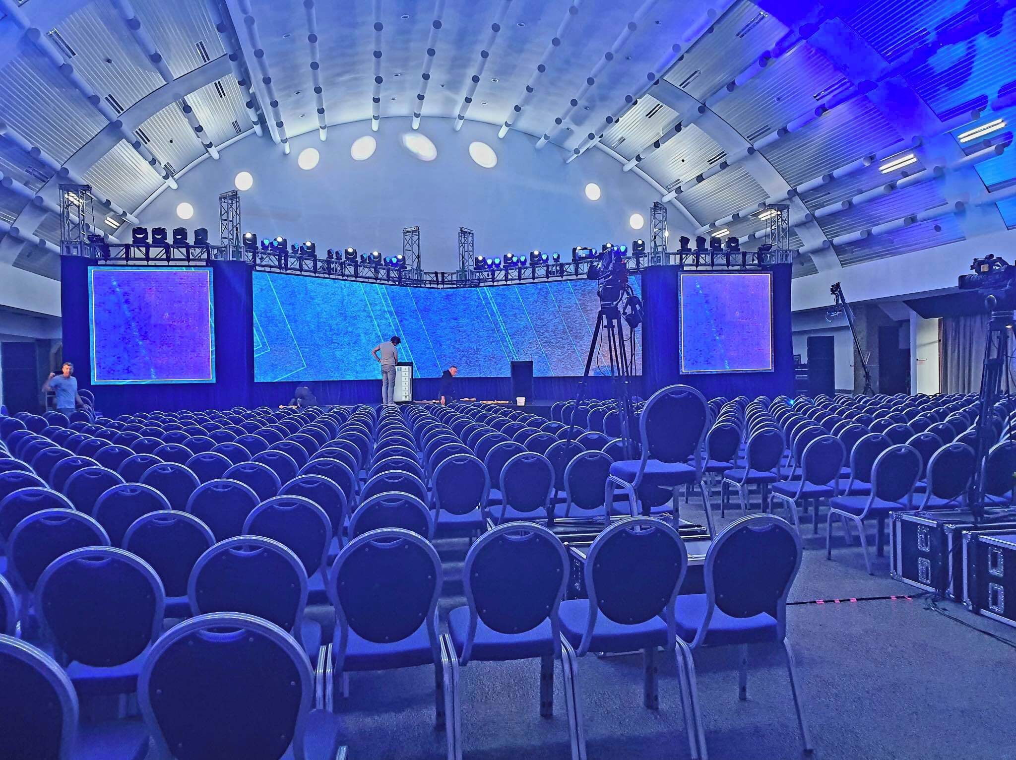 Un auditorio con filas de sillas, un escenario con una pantalla grande y una estructura de soporte colgante.