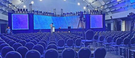 Un auditorio con filas de sillas, un escenario con una pantalla grande y una estructura de soporte colgante.